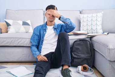 Young hispanic man sitting on the floor studying for university covering eyes with hand, looking serious and sad. sightless, hiding and rejection concept 