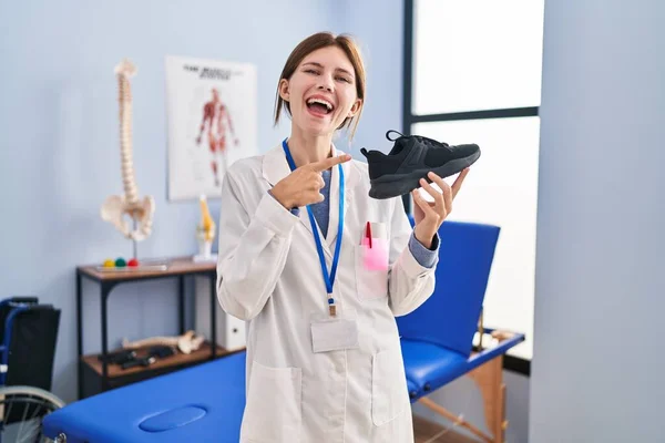 Joven Fisioterapeuta Mujer Sosteniendo Zapatillas Sonriendo Feliz Señalando Con Mano —  Fotos de Stock