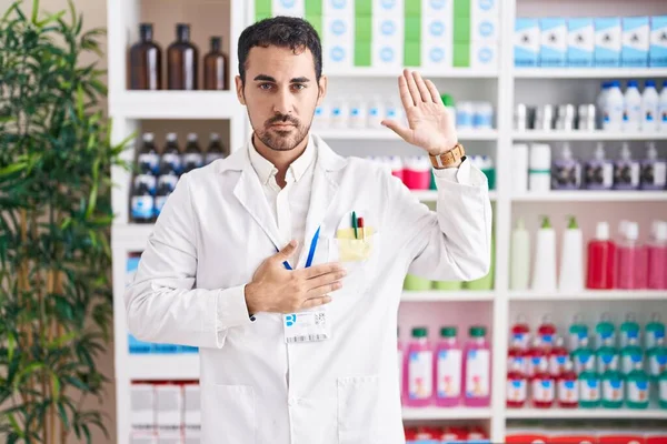 Handsome hispanic man working at pharmacy drugstore swearing with hand on chest and open palm, making a loyalty promise oath