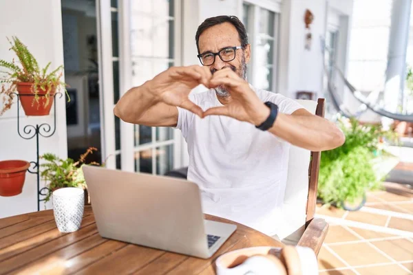 Middle age man using computer laptop at home smiling in love doing heart symbol shape with hands. romantic concept.