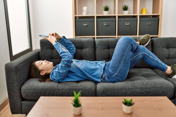 Молодая китаянка с помощью смартфона лежит на диване дома.