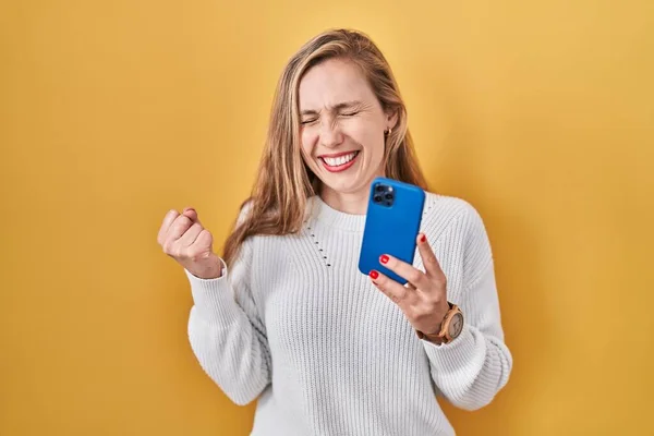 スマートフォンの入力メッセージを使用して若いブロンドの女性は非常に満足し 腕を上げて勝者のジェスチャーを行う興奮し 笑顔と成功のために叫んでいます お祝いのコンセプト — ストック写真