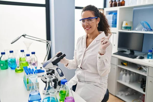Young hispanic woman working at scientist laboratory smiling happy pointing with hand and finger to the side