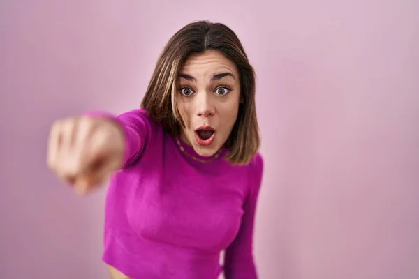 Hispanic woman standing over pink background pointing with finger surprised ahead, open mouth amazed expression, something on the front