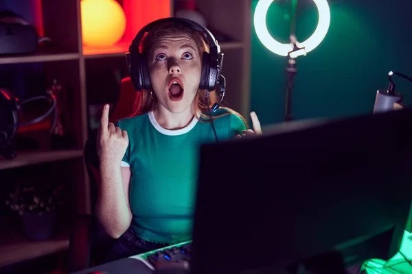 stock image Redhead woman playing video games amazed and surprised looking up and pointing with fingers and raised arms. 