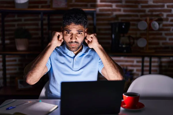 Hispanic man with beard using laptop at night covering ears with fingers with annoyed expression for the noise of loud music. deaf concept.