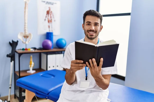 Young Hispanic Man Wearing Physiotherapist Uniform Reading Book Rehab Clinic — Zdjęcie stockowe