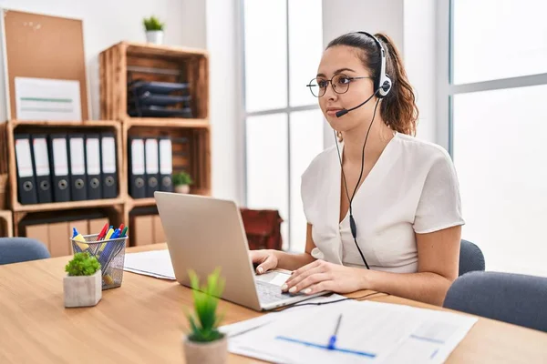 Young hispanic woman wearing call center agent headset at the office looking to side, relax profile pose with natural face with confident smile.