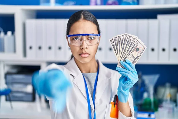 Young brazilian woman working at scientist laboratory holding money pointing with finger to the camera and to you, confident gesture looking serious