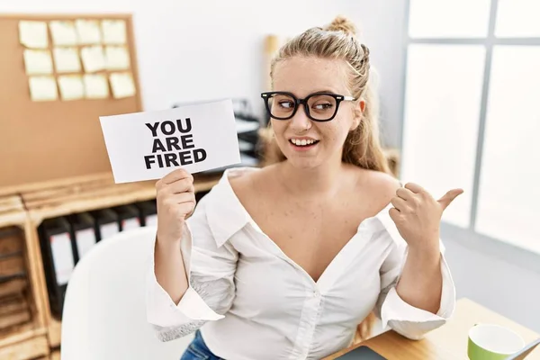 Young caucasian woman holding you are fired banner at the office pointing thumb up to the side smiling happy with open mouth