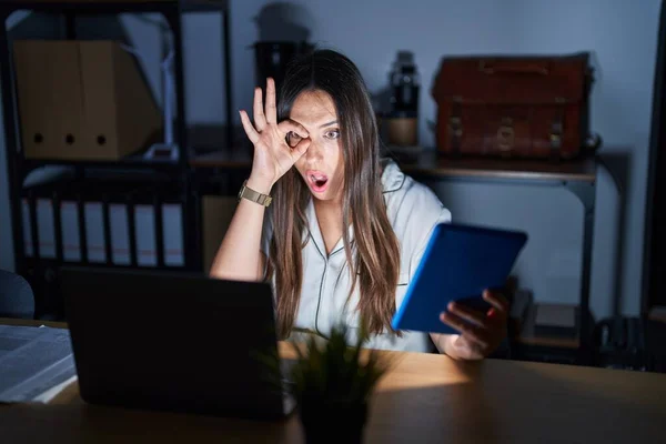 Young brunette woman working at the office at night doing ok gesture shocked with surprised face, eye looking through fingers. unbelieving expression.