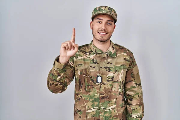 Young hispanic man wearing camouflage army uniform showing and pointing up with finger number one while smiling confident and happy.
