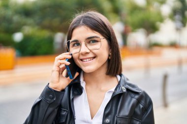 Genç, güzel, İspanyol bir kadın gülümsüyor ve caddede akıllı telefondan konuşuyor.