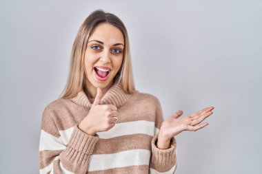 Young blonde woman wearing turtleneck sweater over isolated background showing palm hand and doing ok gesture with thumbs up, smiling happy and cheerful 