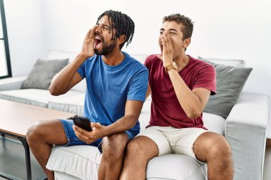 Young hispanic men using smartphone sitting on the sofa at home shouting and screaming loud to side with hand on mouth. communication concept. 