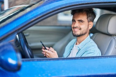 Young hispanic man using smartphone sitting on car at street