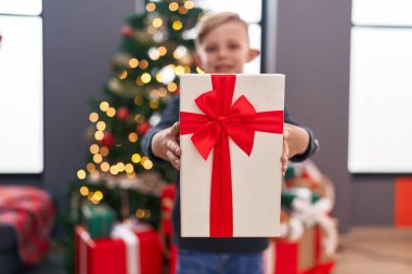 Sevimli çocuk elinde hediyeyle Noel ağacının yanında duruyor.