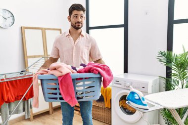 Young man with beard holding laundry basket smiling looking to the side and staring away thinking. 