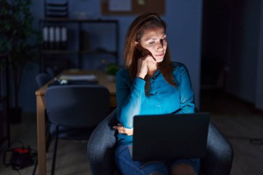 Brunette woman working at the office at night with hand on chin thinking about question, pensive expression. smiling with thoughtful face. doubt concept. 