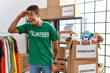 Young handsome hispanic man wearing volunteer t shirt at donations stand very happy and smiling looking far away with hand over head. searching concept. 