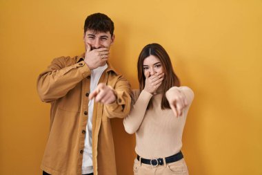 Young hispanic couple standing over yellow background laughing at you, pointing finger to the camera with hand over mouth, shame expression 