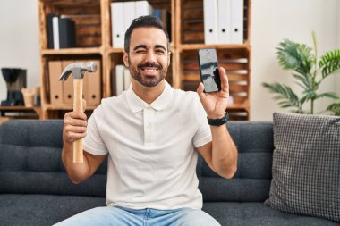 Sakallı, çekiçli ve akıllı telefonlu genç İspanyol adam kameraya seksi bir ifadeyle bakıyor, neşeli ve mutlu bir yüzle.. 