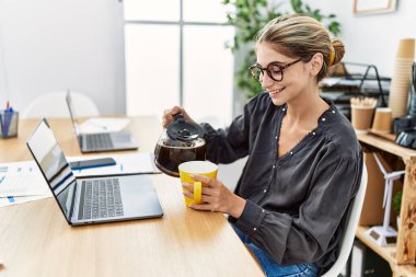 Kahve içen genç beyaz kadın ofiste çalışıyor.