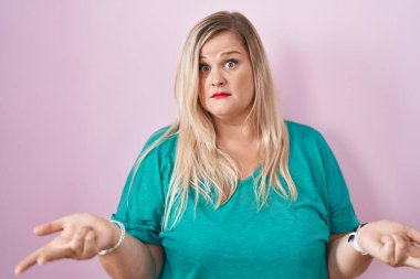 Caucasian plus size woman standing over pink background clueless and confused with open arms, no idea concept. 