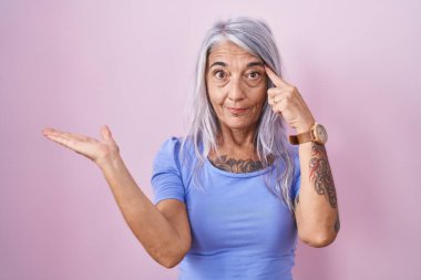 Pembe arka planda duran orta yaşlı kadın avuç içi açık fotokopi alanı ve alnını gösteren dövmelerden rahatsız ve kafası karışık. Bunu bir düşün.. 