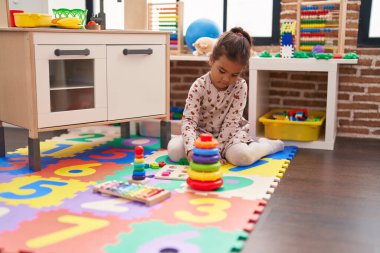 Adorable hispanic girl playing with maths puzzle game sitting on floor at kindergarten
