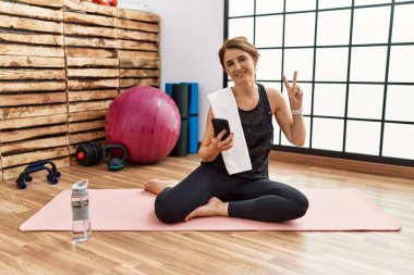 Orta yaşlı bir kadın spor salonundaki antrenman minderinde oturmuş kameraya bakıp parmaklarını zafer işareti yaparken gülümsüyor. İki numara.. 