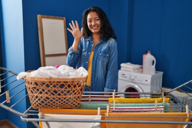 Young asian woman hanging clothes at clothesline showing and pointing up with fingers number five while smiling confident and happy. 