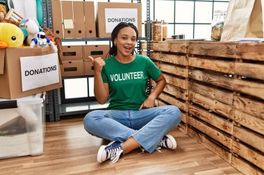 Young african american woman wearing volunteer t shirt at donations stand smiling with happy face looking and pointing to the side with thumb up. 