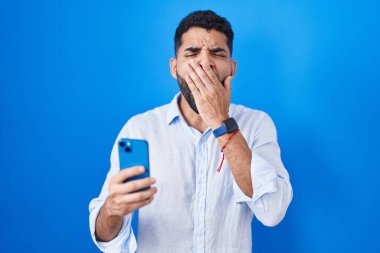 Hispanic man with beard using smartphone typing message bored yawning tired covering mouth with hand. restless and sleepiness. 