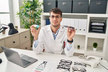 Young hispanic oculist holding glasses and contact lenses relaxed with serious expression on face. simple and natural looking at the camera. 