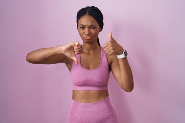 African american woman with braids wearing sportswear over pink background doing thumbs up and down, disagreement and agreement expression. crazy conflict 