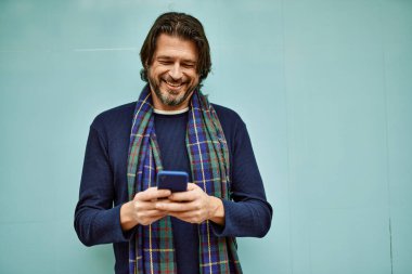 Orta yaşlı yakışıklı adam duvara dayanmış akıllı telefon kullanıyor.