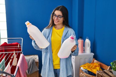 Young hispanic woman smiling confident holding detergent bottles at laundry room