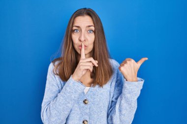 Young woman standing over blue background asking to be quiet with finger on lips pointing with hand to the side. silence and secret concept. 