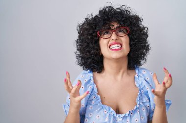 Young brunette woman with curly hair wearing glasses over isolated background crazy and mad shouting and yelling with aggressive expression and arms raised. frustration concept.  clipart