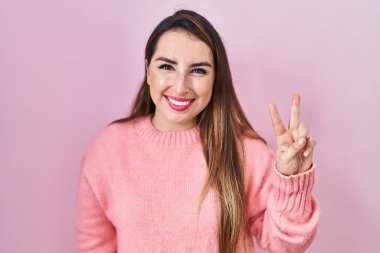 Pembe arka planda duran genç İspanyol kadın iki numarayı gösterip işaret ederken kendinden emin ve mutlu bir şekilde gülümsüyor.. 