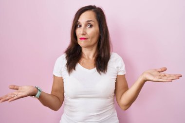 Middle age brunette woman standing over pink background clueless and confused with open arms, no idea concept. 