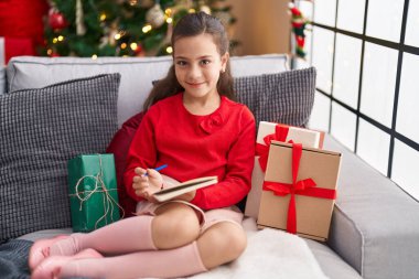 Evdeki noel ağacının yanında, kanepeye oturan sevimli İspanyol kız deftere yazı yazıyor.
