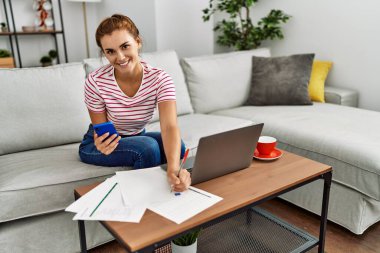 Akıllı telefon ve dizüstü bilgisayar kullanan genç bir kadın evdeki kanepede oturuyor.