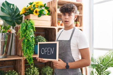 Hispanic teenager working at florist holding open sign scared and amazed with open mouth for surprise, disbelief face 