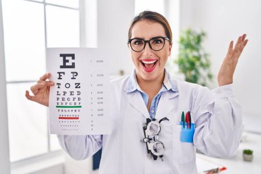 Young hispanic optician woman holding medical exam celebrating victory with happy smile and winner expression with raised hands 
