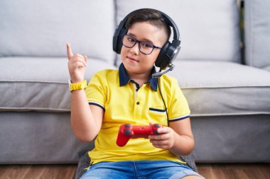 Young hispanic kid playing video game holding controller wearing headphones with a big smile on face, pointing with hand finger to the side looking at the camera. 