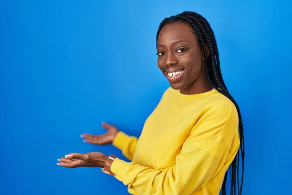 Beautiful black woman standing over blue background inviting to enter smiling natural with open hand