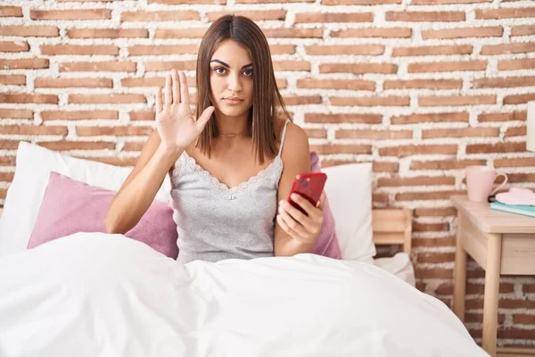 Young hispanic woman using smartphone sitting on the bed at home with open hand doing stop sign with serious and confident expression, defense gesture