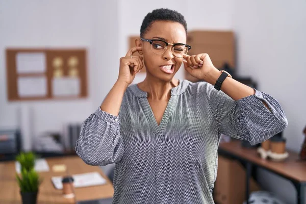 African american woman working at the office wearing glasses covering ears with fingers with annoyed expression for the noise of loud music. deaf concept.